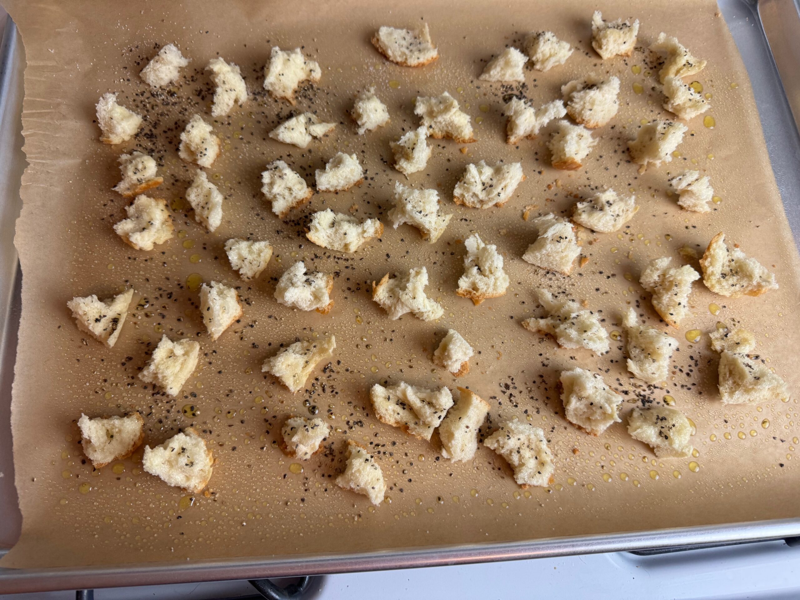 bread sprayed with olive oil and sprinkled with salt and pepper on a baking sheet as a step to making homemade croutons